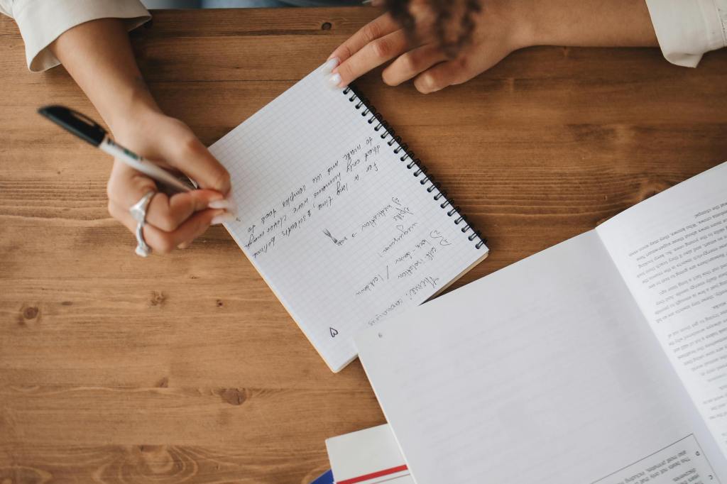 A student wearing a ring writes structured organisational notes in a spiralled notebook during an executive function coaching session.